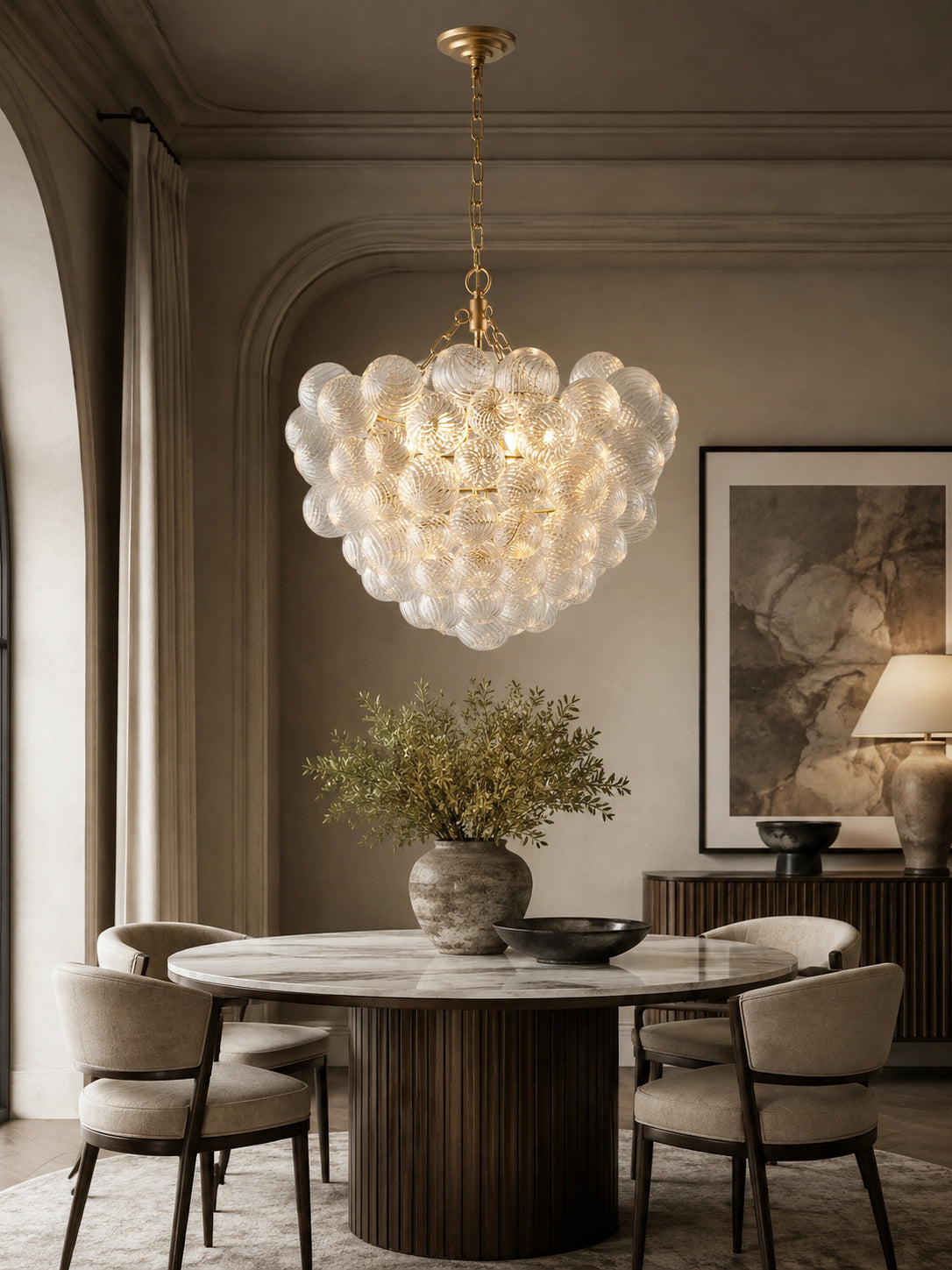 A modern cloud-like chandelier with textured clear glass bubble clusters and warm gold-toned hardware, hanging over a round dining table in a styled interior room.