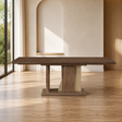 A rectangular wooden dining table with a dark brown top and a sculptural pedestal base in matching wood and light beige stone, set in a modern minimalist room with a wood floor and arched wall niche.