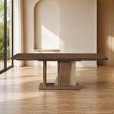 A rectangular wooden dining table with a dark brown top and a sculptural pedestal base in matching wood and light beige stone, set in a modern minimalist room with a wood floor and arched wall niche.
