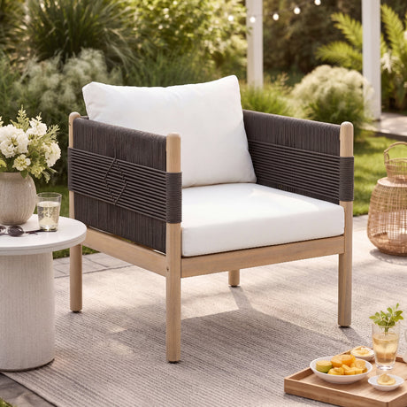A modern outdoor patio armchair with a light wood frame, dark woven armrests, and white cushions, styled on a gray rug in a garden patio setting.