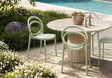 Outdoor patio dining set featuring a round natural stone table and three light sage green molded chairs, set next to a swimming pool with green landscaping.