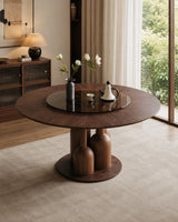 A round dark wood dining table with a glossy black glass top and a sculptural multi-bulb pedestal base, styled in a modern, warm-toned living room.
