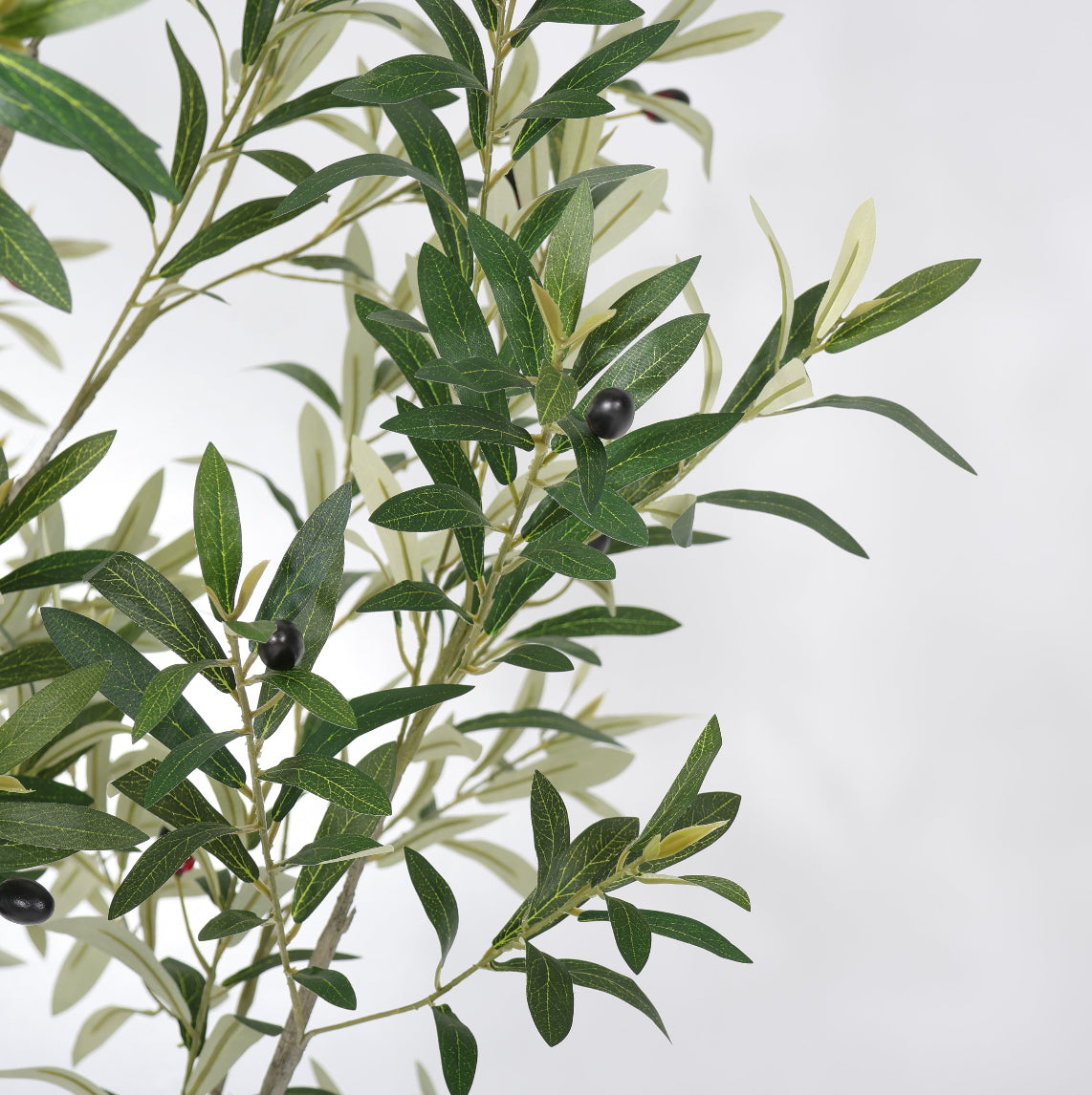 Artificial olive branch with green and silvery-green leaves and small dark olives, photographed against a plain light gray background.