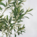 Artificial olive branch with green and silvery-green leaves and small dark olives, photographed against a plain light gray background.
