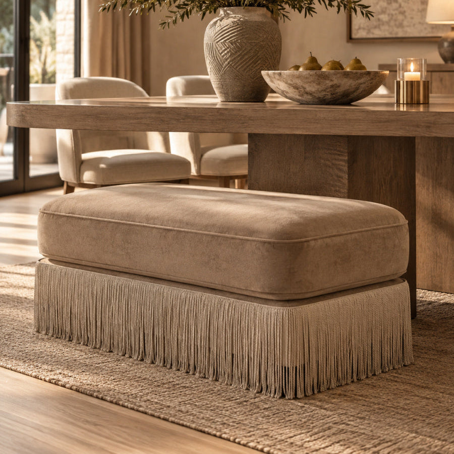 A beige upholstered ottoman with fringe detailing, positioned in a modern, warm-toned dining room setting next to a wooden dining table.