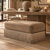 A beige upholstered ottoman with fringe detailing, positioned in a modern, warm-toned dining room setting next to a wooden dining table.