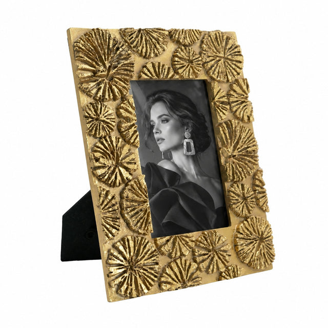 A rectangular gold picture frame with a textured, raised floral or starburst pattern around the border, holding a black-and-white portrait photo. The frame is shown standing on a small black easel against a plain white background.