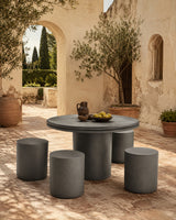 A round outdoor dining table with four matching cylindrical stools in a warm stone courtyard, styled with a small pitcher, bowl, and fruit on top.
